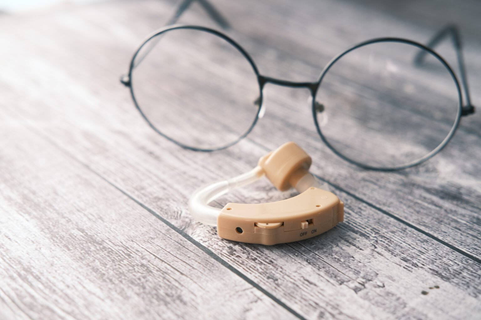 A hearing aid next to a pair of round-framed glasses on a wooden surface, symbolizing audiology equipment.