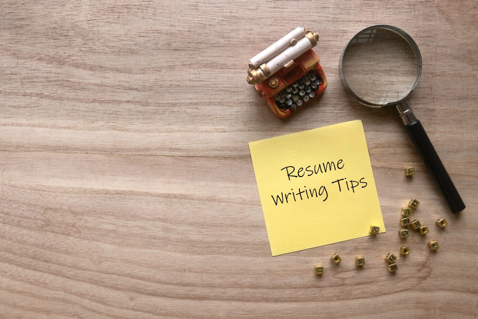 A miniature typewriter, a magnifying glass, and a yellow sticky note with the words 'Resume writing Tips' written on it, placed on a wooden surface. Scattered around are small, golden alphabet letter beads.