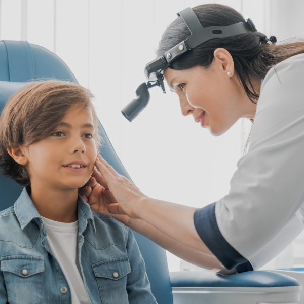 A professional otolaryngologist, equipped with a headlamp, is gently examining a young boy's ear. The boy, dressed in a casual denim shirt, sits comfortably in a specialized ENT chair, looking at the doctor with a calm expression. The environment suggests a modern clinical setting with bright lighting.